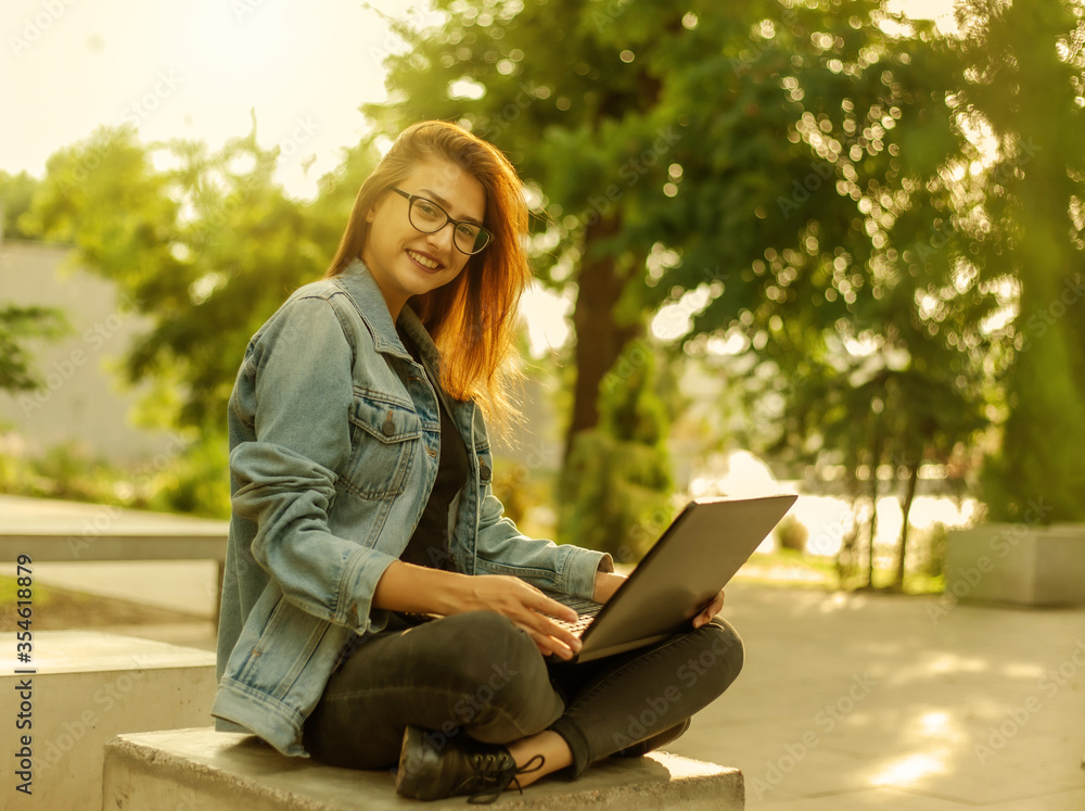 Young modern woman blogger in a denim jacket and glasses holds a laptop in his hands in the park. Online learning, blogging Young modern woman blogger in a denim jacket and glasses holds a laptop in his hands in the park. Online learning, blogging