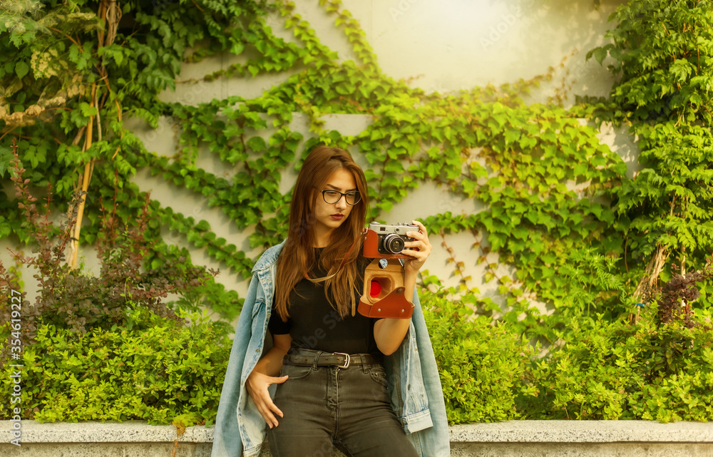 Fototapeta premium Young hipster woman in denim jacket and glasses enjoys film retro camera outdoors