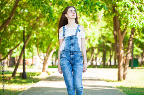 Portrait of a young beautiful girl in blue jeans overalls walking in summer park