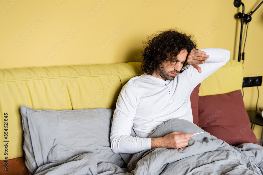 upset man touching forehead while looking at thermometer in bed