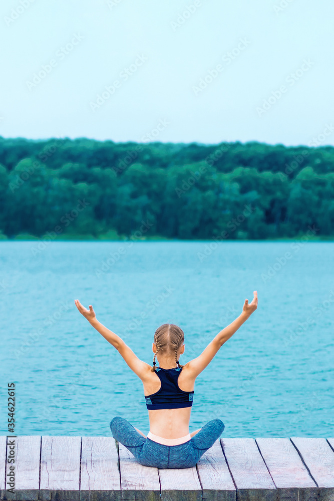 Back view of little girl is doing yoga exercise on grass of the shore of the lake.