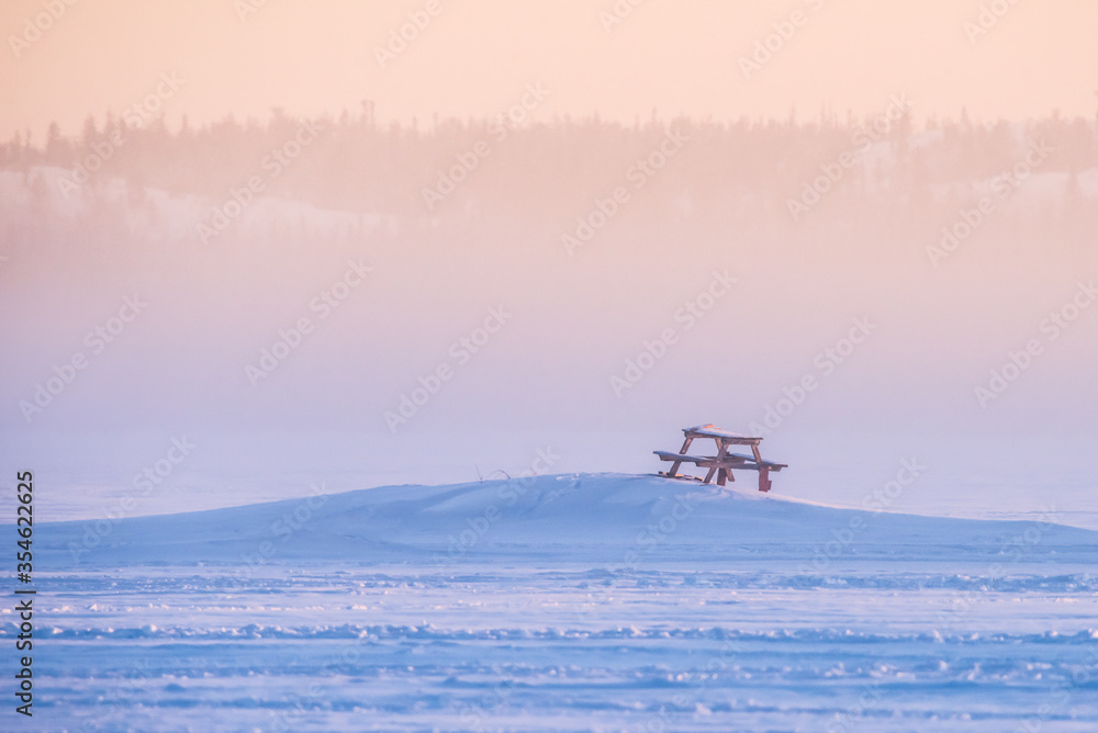 A picnic table surrounded by deep snow at sunrise on a frozen Great Slave Lake in Yellowknife, Northwest Territories, Canada. A picnic table surrounded by deep snow at sunrise on a frozen Great Slave Lake in Yellowknife, Northwest Territories, Canada.