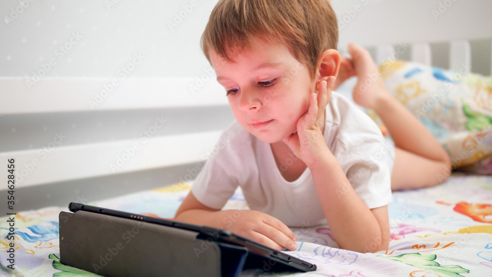 Portrait of cute smiling little boy lying in bed and watching video on digital tablet computer Portrait of cute smiling little boy lying in bed and watching video on digital tablet computer