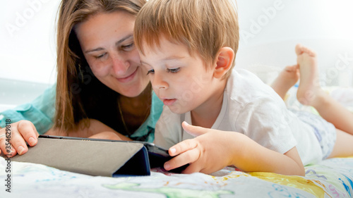 Portrait of cute little boy with young mother playing on digital tablet computer in bed at morning