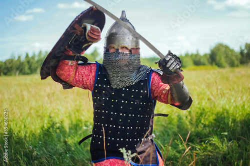 A medieval Central Asian warrior, a nomad in 14th-century armor, stands in a defensive or attacking position with a shield and a saber in his hands in a field against a forest.