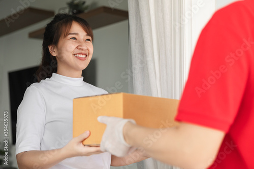 Asian delivery man holding cardboard boxes in medical rubber gloves and mask give to young woman in front of the house. © Suradech