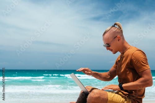 Young guy with long hair by the sea on a sunny day, working using his laptop, working remotely with a business. Blogger, freelancer, work through the Internet, work travel. Summer vacation