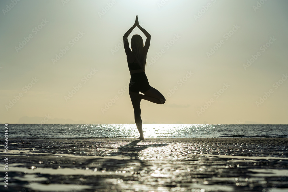Silhouette of woman standing at yoga pose on the tropical beach during sunset. Girl practicing yoga near sea water