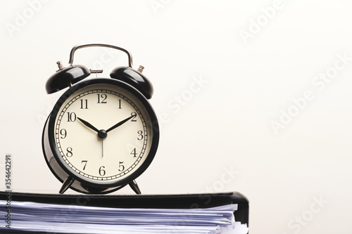 Black vintage alarm clock on paper files stacked in black folder isolated on white wall background.
