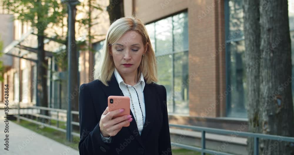 Serious woman in 30s scrolling and looking at smartphone screen while reading news. Beautiful businesswoman using her phone while walking at street near modern office building Serious woman in 30s scrolling and looking at smartphone screen while reading news. Beautiful businesswoman using her phone while walking at street near modern office building