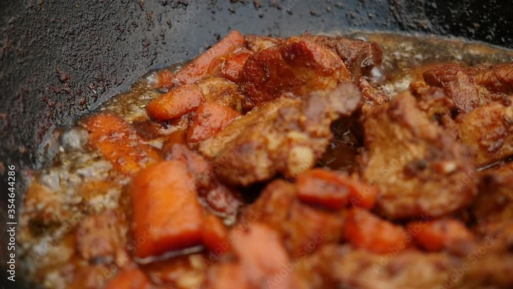 Cooking process, frying pork meat with carrots and onion in large cauldron on open fire. Close up, slow motion Cooking process, frying pork meat with carrots and onion in large cauldron on open fire. Close up, slow motion