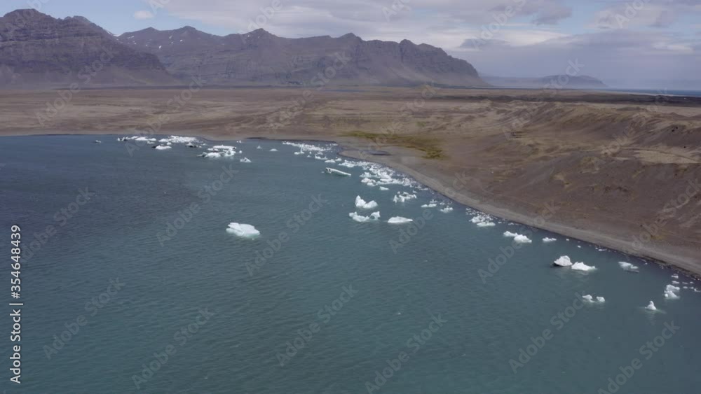 Aerial panning shot of icebergs in lake near sea against cloudy sky, drone descending while flying forward over lagoon - Southwestern Iceland, Iceland Aerial panning shot of icebergs in lake near sea against cloudy sky, drone descending while flying forward over lagoon - Southwestern Iceland, Iceland