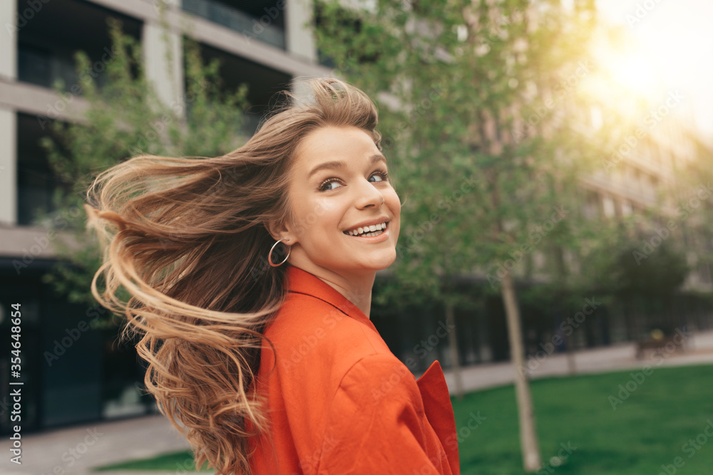 Beautiful gorgeous young woman wave her hair and look back with smile. Picture in action. Stylish trendy girl walking outside on street at urban building. Posing in orange jacket.