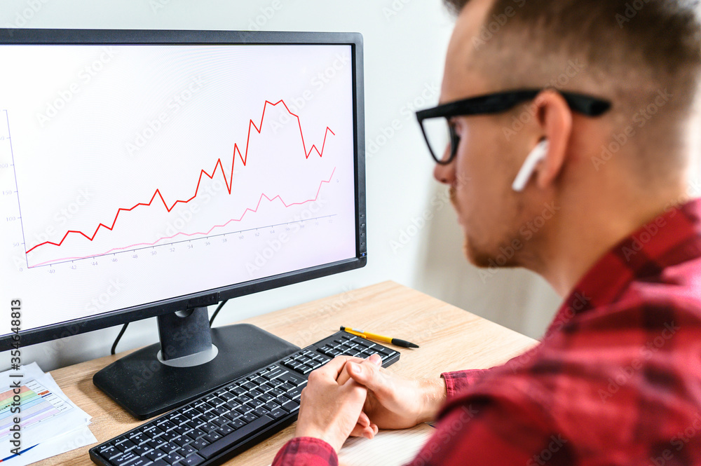 A young serious man looks at the PC screen with graphs on it, he sits at the office desk with hands on it, he is in glasses and headphones, a side view from his back A young serious man looks at the PC screen with graphs on it, he sits at the office desk with hands on it, he is in glasses and headphones, a side view from his back
