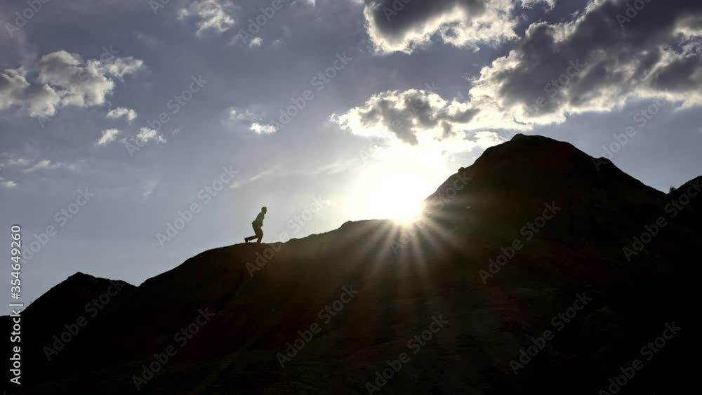 The general plan of the mountain with different peaks, on the steep slope of which the silhouette of a businessman rises, he overcomes the obstacle and, being at the top, raises his hands in an The general plan of the mountain with different peaks, on the steep slope of which the silhouette of a businessman rises, he overcomes the obstacle and, being at the top, raises his hands in an