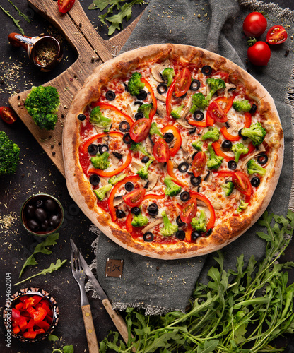 Vegetarian pizza with broccoli, red pepper, tomato and black olives on the wooden cupboard and some ingredients and spices around 