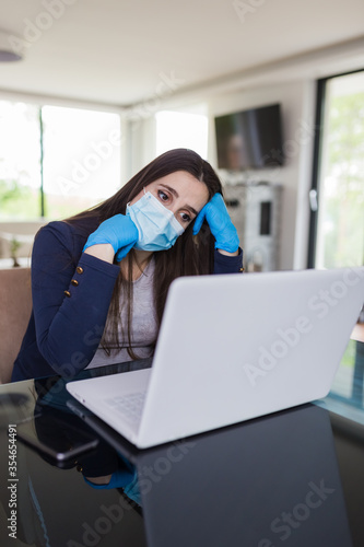 Young sad businesswoman working on laptop from home, wearing protective mask and gloves. Coronavirus quarantine and business problems concept.