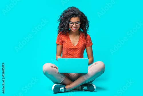 Beautiful young smiling woman in casual outfit and trendy eyeglasses sitting isolated on bright colored blue background and working on her laptop photo