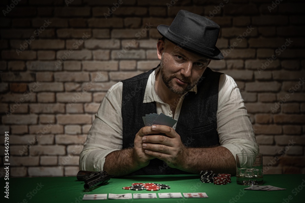 Poker player sitting behind the poker table with cards and playing chips - poker in a dark back room with a brick wall Poker player sitting behind the poker table with cards and playing chips - poker in a dark back room with a brick wall
