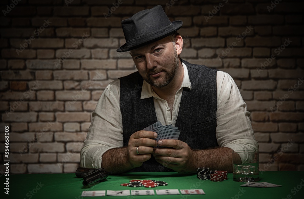 Poker player sitting behind the poker table with cards and playing chips - poker in a dark back room with a brick wall Poker player sitting behind the poker table with cards and playing chips - poker in a dark back room with a brick wall