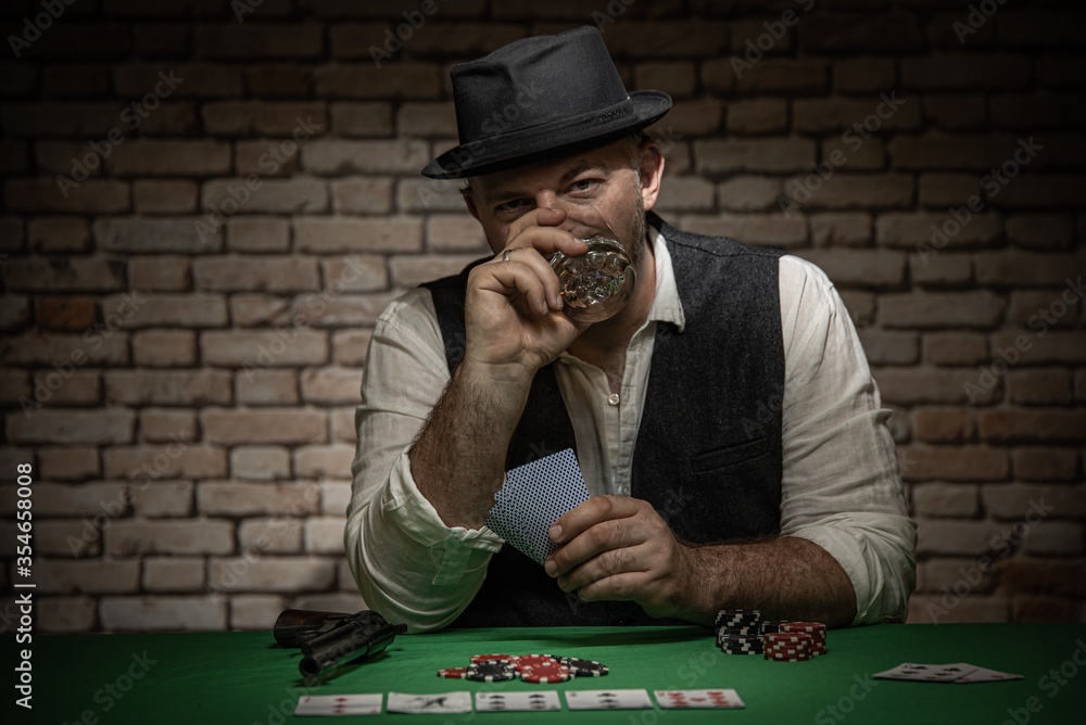 Poker player sitting behind the poker table with cards and playing chips - poker in a dark back room with a brick wall Poker player sitting behind the poker table with cards and playing chips - poker in a dark back room with a brick wall