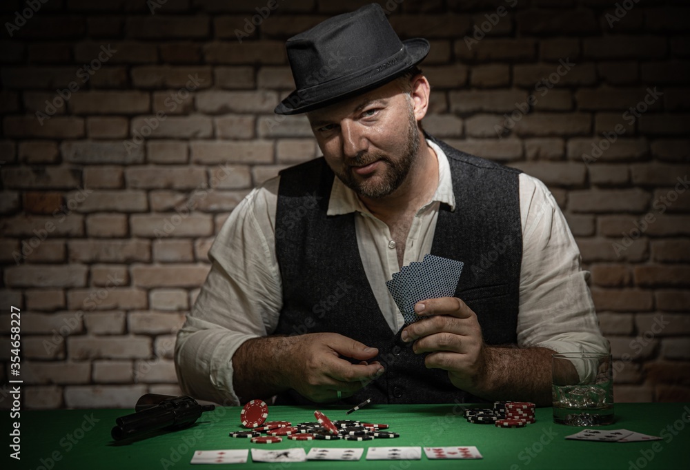 Poker player sitting behind the poker table with cards and playing chips - poker in a dark back room with a brick wall Poker player sitting behind the poker table with cards and playing chips - poker in a dark back room with a brick wall