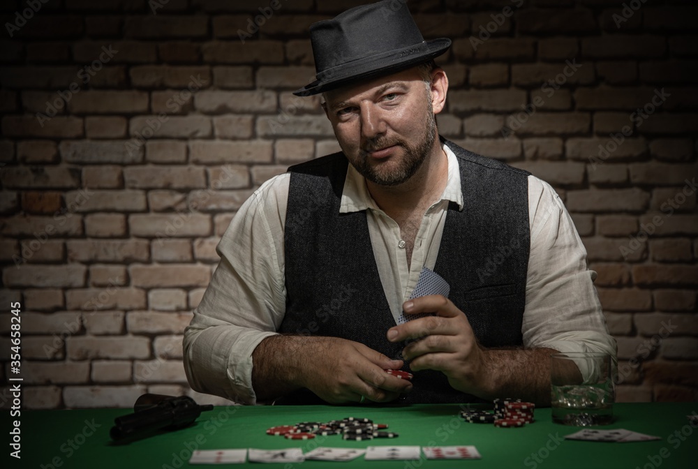 Poker player sitting behind the poker table with cards and playing chips - poker in a dark back room with a brick wall Poker player sitting behind the poker table with cards and playing chips - poker in a dark back room with a brick wall