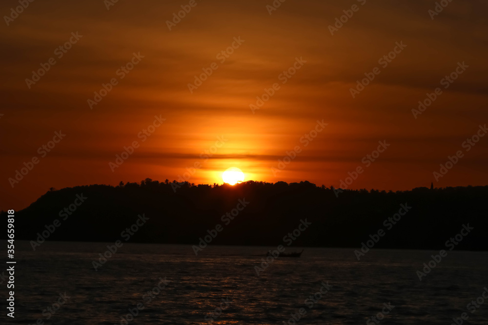 Sunset with the orange sky behind the mountain surrounded by the sea with the dark silhouette 