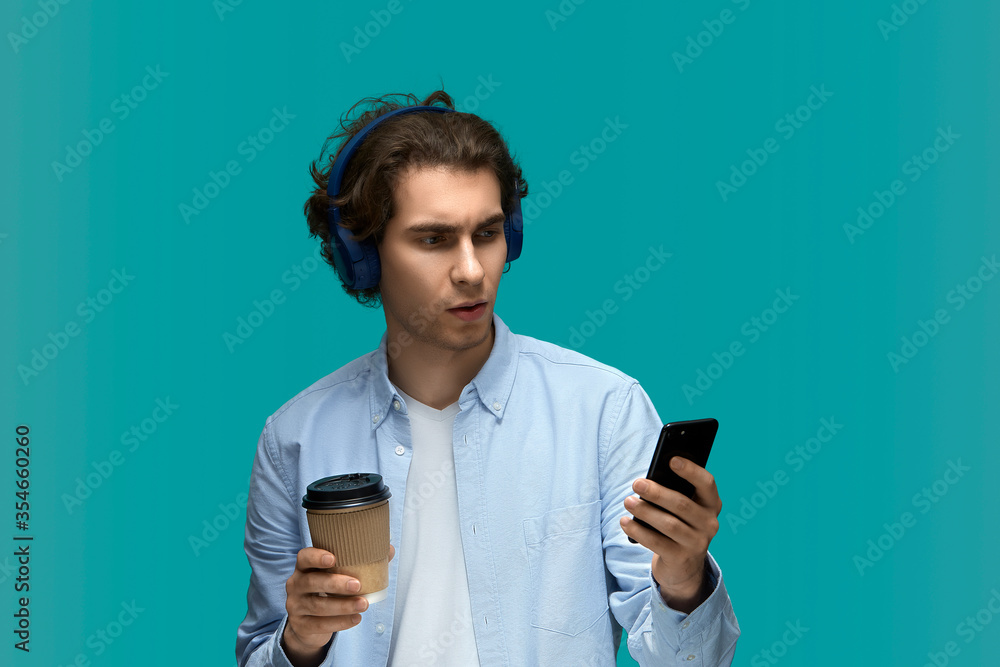 Morning surprise. Portrait of a young beautiful man wearing white t-shirt and blue shirt in blue headphones holding disposable cup with drink and looking at mobile phone with wide open eyes Morning surprise. Portrait of a young beautiful man wearing white t-shirt and blue shirt in blue headphones holding disposable cup with drink and looking at mobile phone with wide open eyes