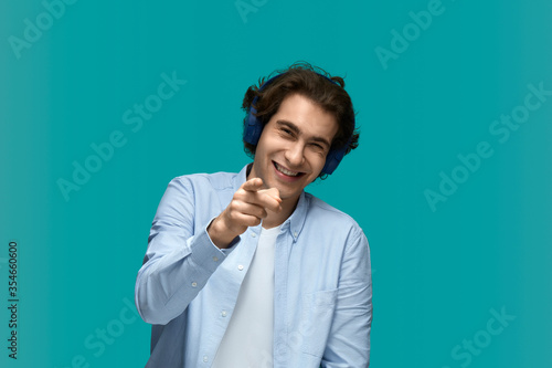 make fun of. Portrait of a young beautiful man wearing white t-shirt and blue shirt in blue headphones laughs, pointing by finger and looking at camera