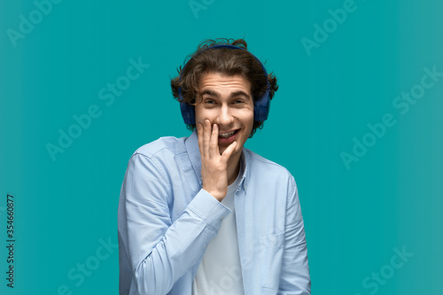 Very funny shame. Portrait of a young beautiful man wearing white t-shirt and blue shirt in blue headphones holds palm in face laugh and looking at camera