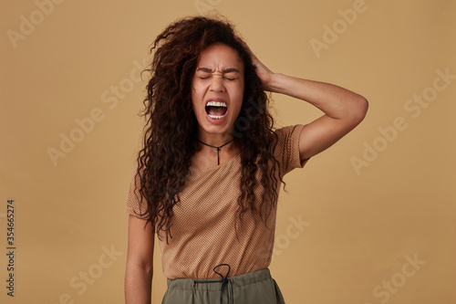 Stressed young brown haired curly dark skinned lady frowning her face while screaming and raising emotionally hand to her head while posing over beige background