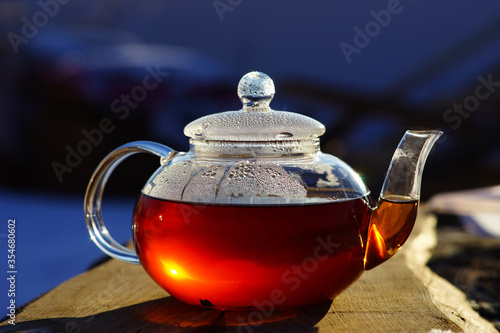 Glass teapot with black tea. The sun is reflected in the teapot. Boiling water, hot tea. Drops on a glass teapot. Tea on the street in the village.