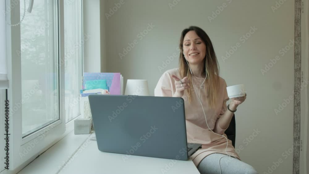 Happy caucasian young woman wear headset communicating by conference call speak looking at computer at home office, video chat job interview or distance work, drink tea Happy caucasian young woman wear headset communicating by conference call speak looking at computer at home office, video chat job interview or distance work, drink tea
