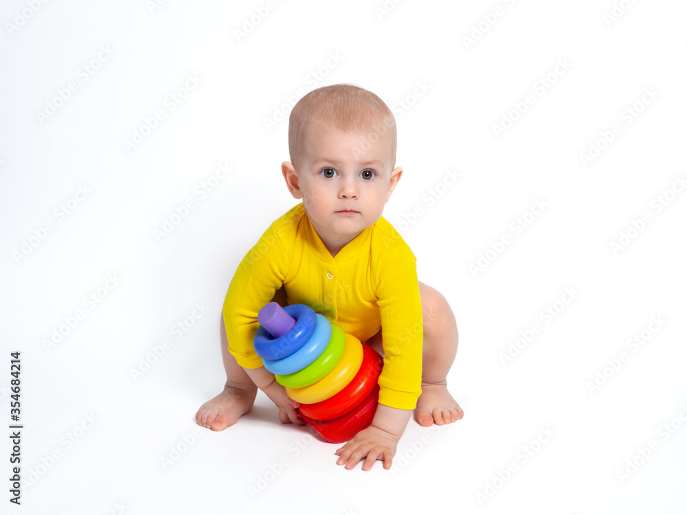 The baby is squatting with a pyramid. Carefully looks into the frame. The little girl is wearing a bright yellow bodysuit on a white background. Toys for babies. The baby is squatting with a pyramid. Carefully looks into the frame. The little girl is wearing a bright yellow bodysuit on a white background. Toys for babies.