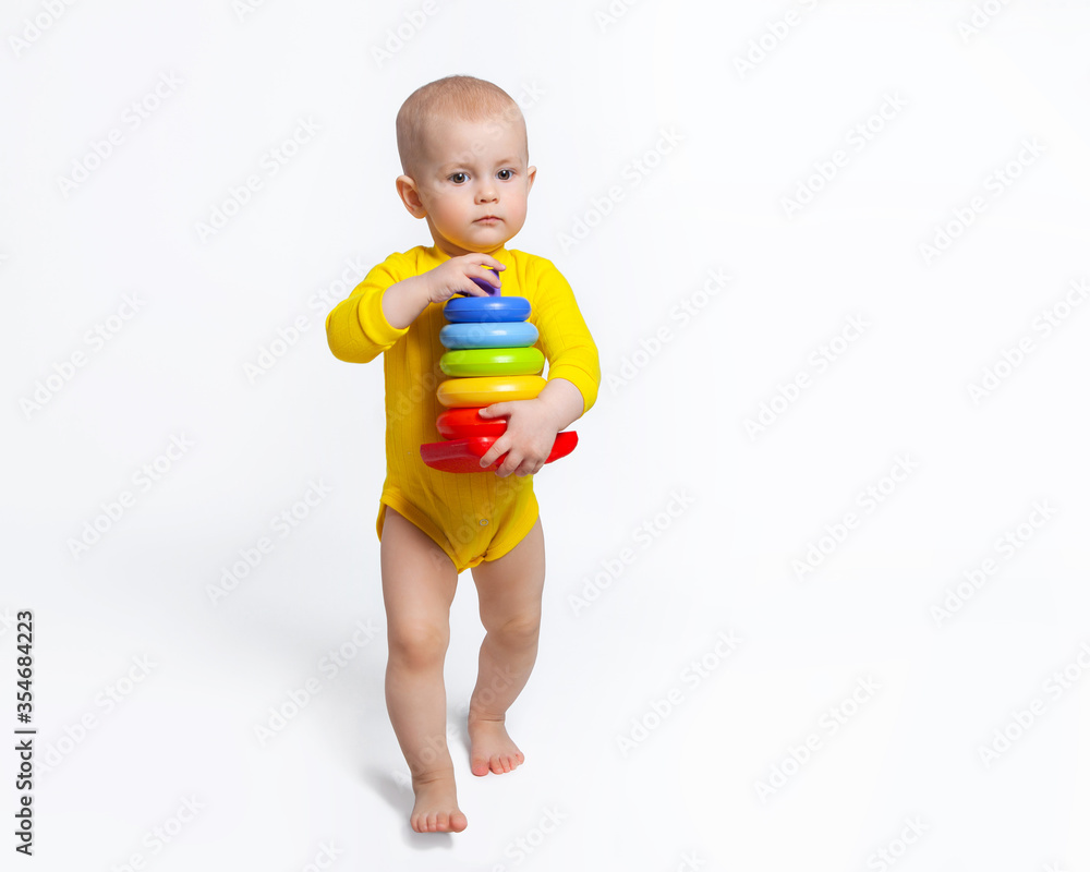 The baby is carrying a pyramid in her hands, walking barefoot on a white background. Baby's first steps. Games and toys for kids. Education and development of children. The baby is carrying a pyramid in her hands, walking barefoot on a white background. Baby's first steps. Games and toys for kids. Education and development of children.