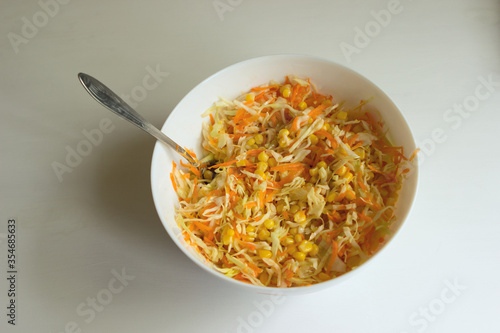 Vegetarian diet low-calorie raw food salad of white cabbage, carrots and sweet corn in a white deep bowl with a spoon on a white table background.