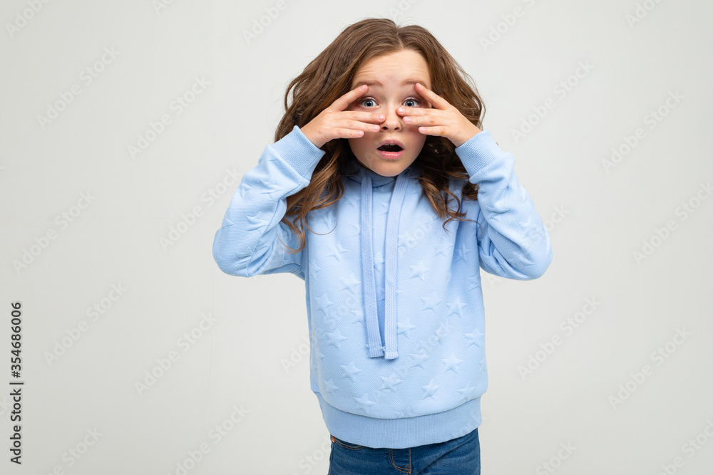 Caucasian teenage girl in a blue hoodie covered her eyes with her hands on a white background with copy space Caucasian teenage girl in a blue hoodie covered her eyes with her hands on a white background with copy space