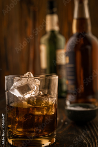 A glass of whiskey with ice, in the background are bottles on a wooden table of a bar counter, shallow depth of field, selective focus. The concept of alcoholic drinks in a roadside bar.