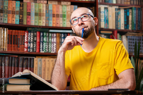 A bald, bearded man with glasses raised a pen to his mouth thoughtfully as he sat in front of his books. There are bookshelves in the background. Concept of distance learning and education