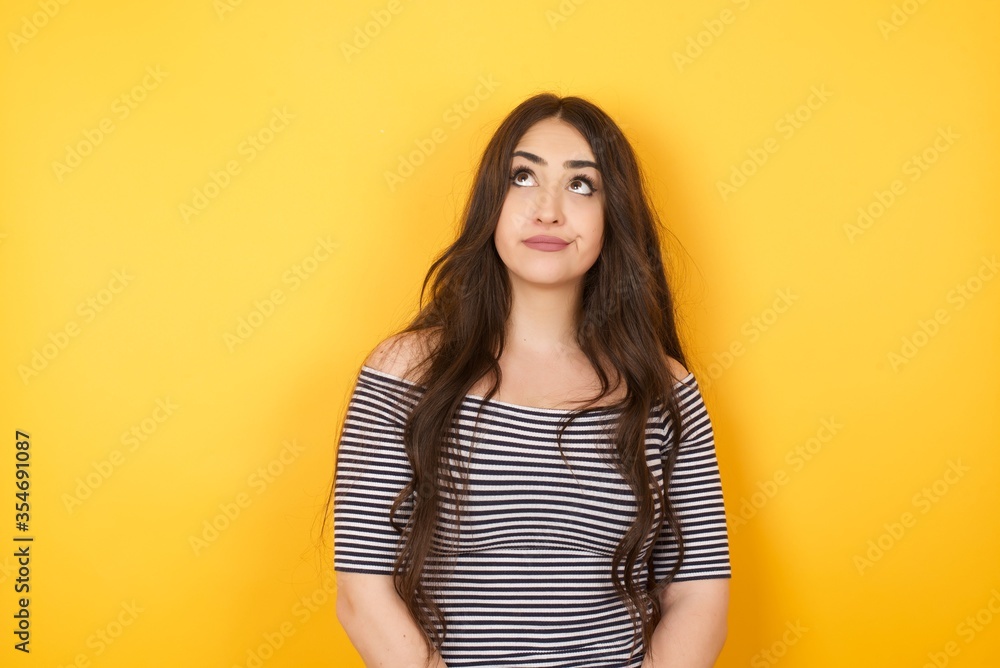 Troublesome puzzled Caucasian woman frowns face, bites lip, raises eyebrows, looks up, has problems, dressed in fashionable clothes, isolated over gray background, has regret look. Troublesome puzzled Caucasian woman frowns face, bites lip, raises eyebrows, looks up, has problems, dressed in fashionable clothes, isolated over gray background, has regret look.