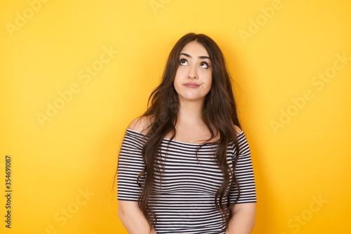 Troublesome puzzled Caucasian woman frowns face, bites lip, raises eyebrows, looks up, has problems, dressed in fashionable clothes, isolated over gray background, has regret look.