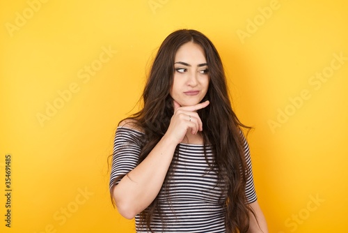 Dreamy  female with pleasant expression, wearing casual clothes, looks sideways, keeps hand under chin, thinks about something pleasant, poses against gray background.