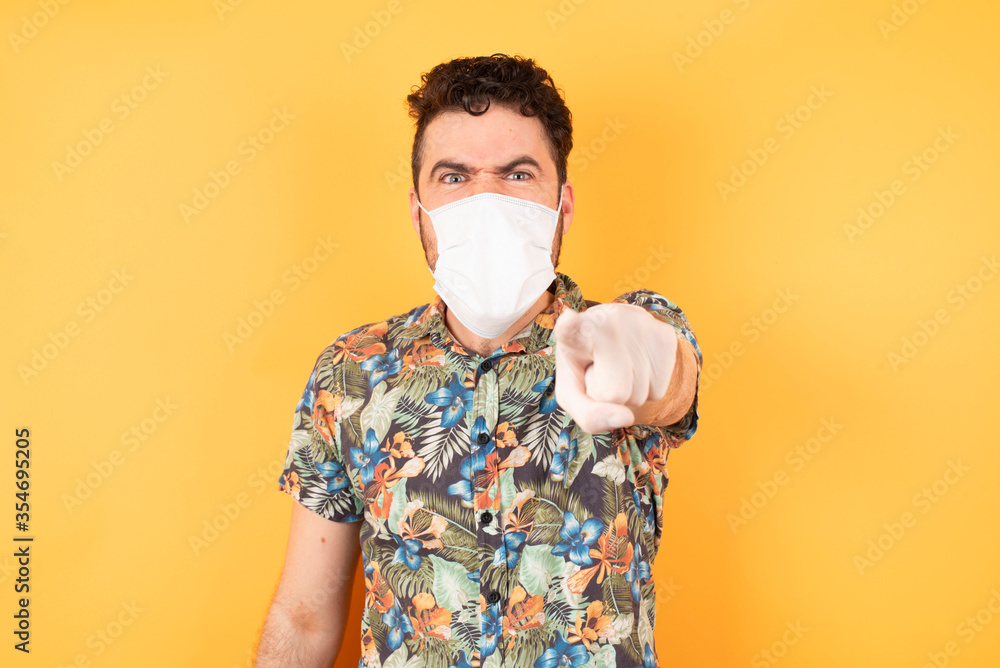 Young businessman wearing hawaiian shirt and medical mask standing over isolated yellow background pointing displeased and frustrated to the camera, angry and furious ready to fight with you. Young businessman wearing hawaiian shirt and medical mask standing over isolated yellow background pointing displeased and frustrated to the camera, angry and furious ready to fight with you.