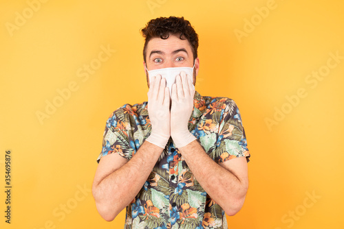 Young  handsome male giggles joyfully, covers mouth, has fun alone, stands against yellow background, has natural laughter, hears positive story or funny anecdote. Hawaiian shirt and medical mask © Roquillo