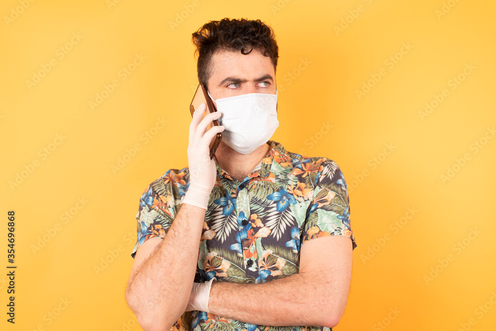 Sad brunette caucasian man in hawaiian shirt and medical mask talking on smartphone on yellow background. Communication concept. Sad brunette caucasian man in hawaiian shirt and medical mask talking on smartphone on yellow background. Communication concept.