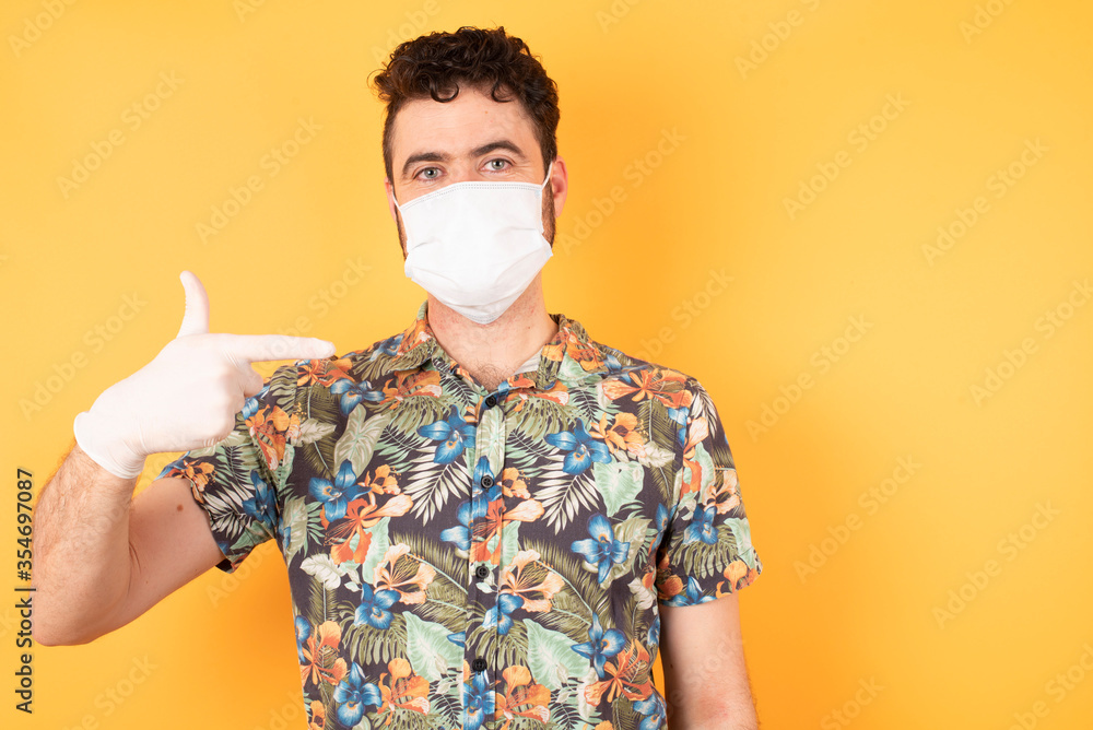 Young handsome Cucasian businessman wearing hawaiian shirt and medical mask standing against yellow background pointing up with fingers number eight in Chinese sign language BÄ. Young handsome Cucasian businessman wearing hawaiian shirt and medical mask standing against yellow background pointing up with fingers number eight in Chinese sign language BÄ.