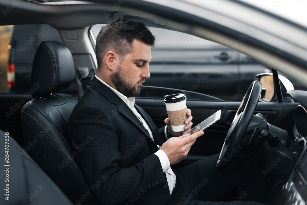 Handsome caucasian businessman driver inside in vehicle with coffee and gadget and check mail and talking with partners Handsome caucasian businessman driver inside in vehicle with coffee and gadget and check mail and talking with partners