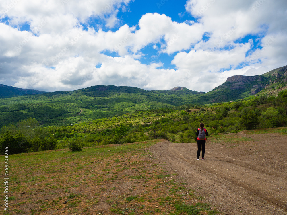 A young girl in a red tank top with a gray backpack in the mountains with green trees and clouds.