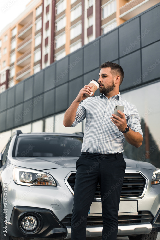 Young handsome man with smartphone and car keys drink black coffee and wait for friend near city parking Young handsome man with smartphone and car keys drink black coffee and wait for friend near city parking